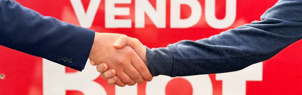 Gros plan sur une poignée de mains entre deux hommes, devant un panneau "Vendu" du groupe Blot.